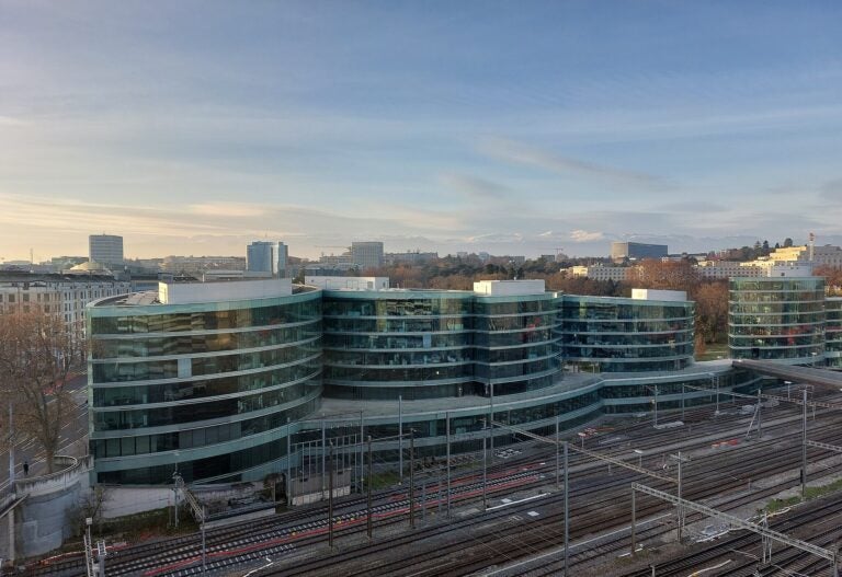 Modern glass buildings of the Geneva Graduate Institute beside railway tracks, with Geneva’s skyline and distant mountains in the background.