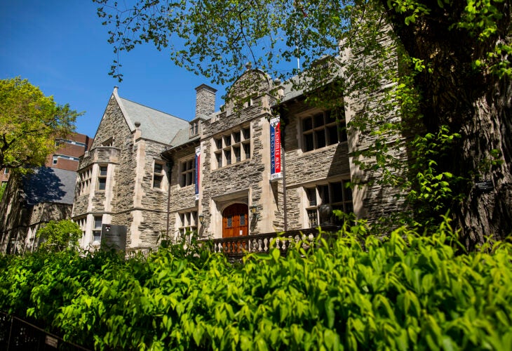 Houston Hall at the University of Pennsylvania, a historic stone building with arched wooden doors framed by "welcome new students" banners, and leafy greenery under a blue sky.