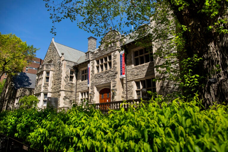 Houston Hall at the University of Pennsylvania, a historic stone building with arched wooden doors framed by "welcome new students" banners, and leafy greenery under a blue sky.