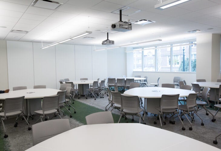 Several round tables surrounded by chairs in classroom with projectors on the ceiling.